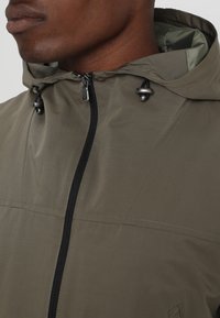 Närbild av en person som bär en olivgrön hoodiejacka med svart dragkedja och justerbara snoddar på huvan.