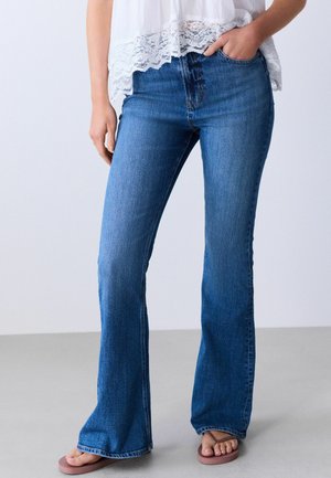 Frau trägt hoch taillierte blaue Bootcut-Jeans, weißes Top mit Spitzenbesatz und pinke Flip-Flops, eine Hand in der Tasche.