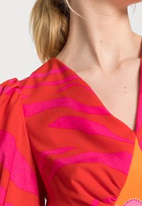 Blouse rouge avec des rayures zèbres rose vif, mettant en avant un décolleté en V et des manches courtes bouffantes. Tissu lisse avec une texture légère.