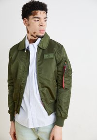 Olivno zelena bomber jakna z gladko teksturo, rebrastimi manšetami in ovratnikom. Ima našitek in žep z zadrgo z rdečim potegom. Oblečena preko svetlo modre srajce z ozkimi črtami.