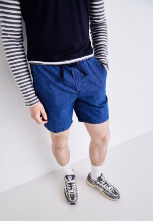 Personne portant un short en jean bleu, un t-shirt à manches longues rayé, des chaussettes blanches et des baskets grises, debout contre un mur blanc.