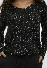 Maglione nero con scollo a barca, ricoperto di paillettes per texture e brillantezza, abbinato a pantaloni neri larghi.