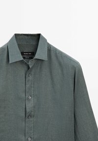 Chemise longue en lin vert à boutons, avec col et étiquette noire à l’intérieur de l’encolure.
