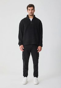 Pullover in pile nero con collo a mezza zip e superficie testurizzata, abbinato a pantaloni cargo neri con tasche laterali e polsini aderenti.