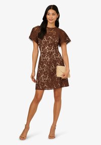 Robe en dentelle marron avec des manches courtes et volants, et une jupe évasée. Le mannequin tient une pochette beige clair et porte des sandales à talons nude.