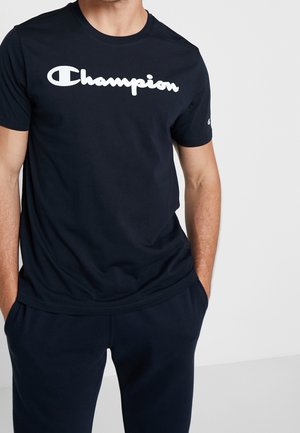 Mężczyzna ubrany w czarną koszulkę Champion i dopasowane czarne spodnie, z rękami w kieszeniach, stojący na tle jasnoszarego tła.