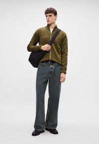 Olive green zip-up knit sweater with patterned texture, paired with loose-fit dark denim jeans. Model carries a black shoulder bag.