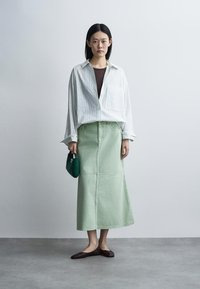 Femme portant une chemise blanche oversize, un top marron, une jupe midi vert menthe, des ballerines marron, tenant un sac à main vert foncé sur un fond clair uni.