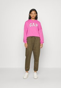 Pink hoodie med "GAP" logo, parret med olivengrønne joggerbukser. Begge items har en afslappet pasform og et afslappet, komfortabelt design. Hvide sneakers.