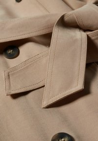Zbliżenie na beżowy materiał z paskiem nawiązuje do fasonu, guzikiem i skośnym prążkowanym wykończeniem w dopasowanym kolorze. Guzik ma czterodziurkowy design.