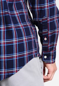 Camisa de cuadros azul marino con detalles en rojo y blanco, puños con botones y dobladillo curvado en los costados. Confeccionada con tela tejida que presenta un patrón general de cuadros.