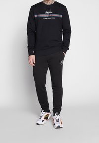 Felpa nera con logo e strisce blu/rosse, abbinata a pantaloni sportivi neri con logo. Indossata con sneakers bianche e gialle.