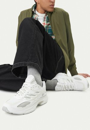 Persona sentada con zapatillas blancas, calcetines grises, pantalones negros, chaqueta verde y camisa de cuadros verde, sobre un fondo liso y claro.