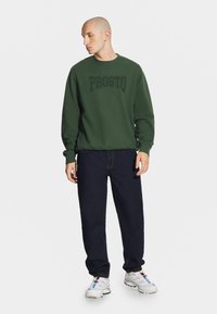 Zielona bluza z kapturem z logo "PROSTO", w połączeniu z ciemnoniebieskimi dżinsami i białymi butami sportowymi, z wielokolorowymi akcentami i grubą podeszwą.