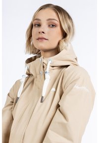Chaqueta impermeable beige con capucha, con cordones blancos y detalles metálicos. Logotipo impreso en la manga izquierda. Textura suave.