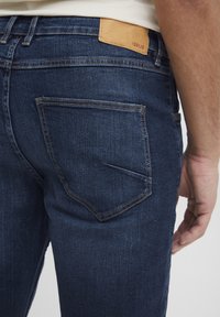 Jeans de mezclilla azul oscuro con una textura suave, que cuentan con un parche de cuero etiquetado como "ISOLID" en la cinturilla y dos bolsillos traseros.