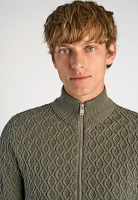 Grön stickad zip-up tröja med hög krage, med ett diamantt mönster och diskreta ribbade detaljer längs kragen och dragkedjan.