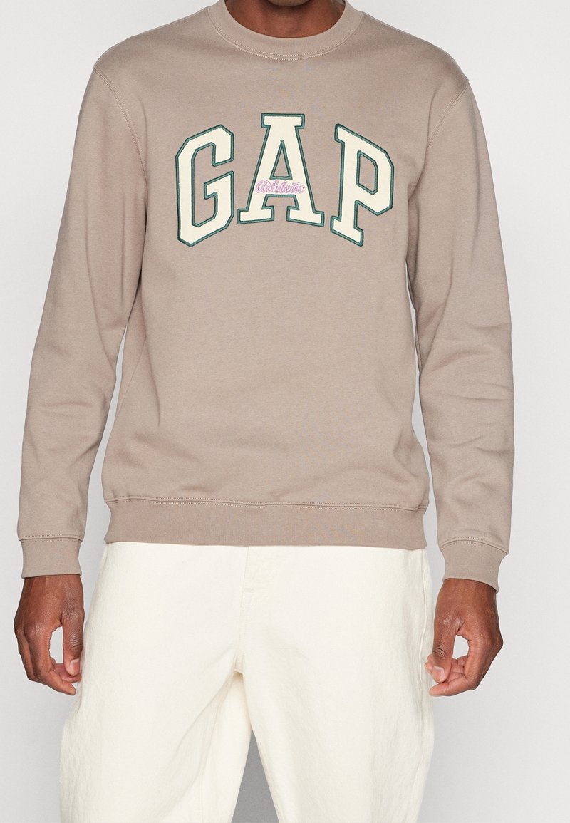 Homme portant un sweat-shirt beige avec de grandes lettres GAP crème et vertes et un texte "Athletic" rose clair, assorti à un pantalon écru.