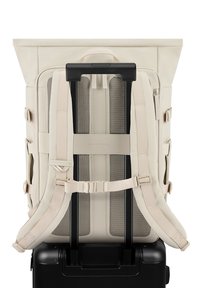 Bežový batoh s nastavitelnými popruhy, síťovinovým polstrováním a designem s rolovacím víkem, umístěný na černém kufru na kolečkách, který ukazuje hladké textury.