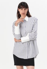 Chemise rayée en blanc et marine avec un col classique, un boutonnage à l'avant et des manches longues avec des poignets blancs contrastants ; ourlet courbé sur les côtés.