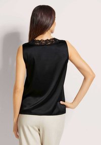 Zwarte mouwloze blouse met een kanten rand bij de halslijn, met een gladde satijnen afwerking en een relaxte pasvorm. Gecombineerd met lichtgekleurde broek.