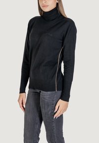 Pullover noir à col roulé avec une texture côtelée et une coupe slim, arborant une subtile couture latérale beige. Associé à un jean gris foncé.