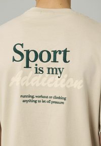 T-shirt en coton beige avec un col rond. Présente un texte brodé vert : « Le sport est ma dépendance » et un texte plus petit en dessous en vert foncé.
