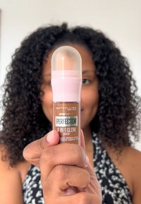 Maybelline Instant Anti-Age Perfector 4-en-1 Glow Make-up dans un flacon en verre avec un bouchon rose arrondi et un liquide bronzé visible à l'intérieur.