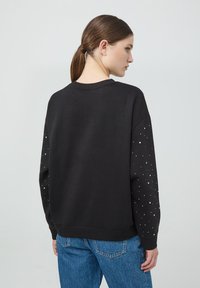 Czarna bluza z okrągłym dekoltem i długimi rękawami. Rękawy zdobione złotymi ćwiekami. Materiał wydaje się miękki; luźny krój z prążkowanym wykończeniem.