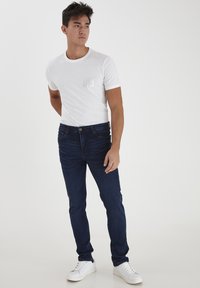 Camiseta blanca de cuello redondo con un pequeño bolsillo texturizado, combinada con jeans ajustados de color azul oscuro y zapatillas blancas.