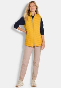 Gepolsterte gelbe Weste mit hohem Kragen, durchgehendem Reißverschluss und Seitentaschen, kombiniert mit einem marineblauen Langarmshirt und hellbeigen Hosen.