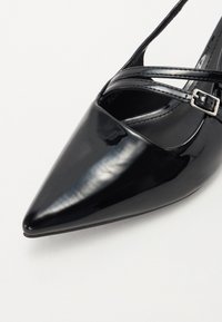 Chaussure à bout pointu en cuir verni noir avec des brides croisées à la cheville et un petit détail de boucle. Texture lisse avec une finition brillante.