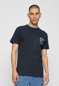 Mørk navy blå bomulds t-shirt med korte ærmer, der har hvid tekst "ALDRIG TIL TID" på venstre side i en afslappet skrifttype.
