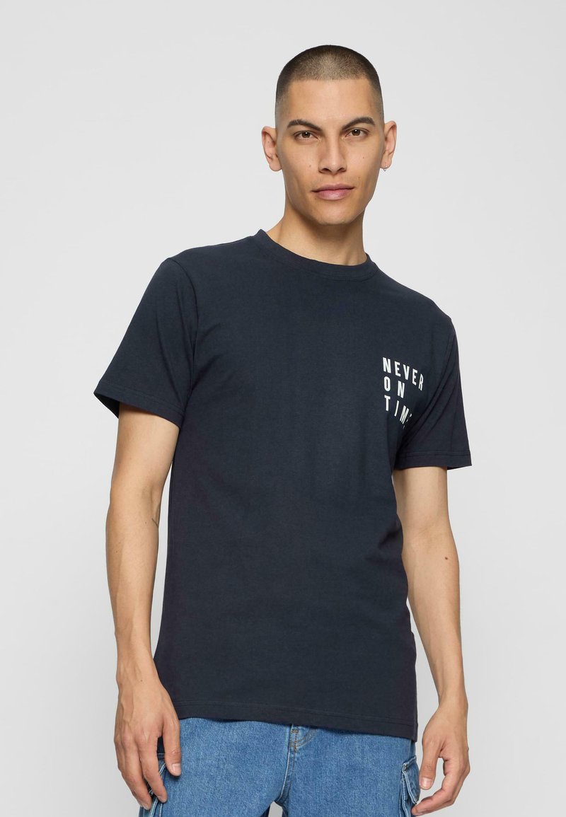 Mørk navy blå bomulds t-shirt med korte ærmer, der har hvid tekst "ALDRIG TIL TID" på venstre side i en afslappet skrifttype.