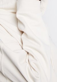 Krämfärgad sweatshirt som visar strukturerad tyg, ribbade muddar och lager-på-lager drapering; har en synlig dragkedja och huva.