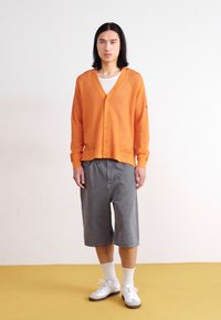 Orange Strickjacke mit V-Ausschnitt und Knopfleiste, kombiniert mit grauen Shorts. Das Model trägt weiße Socken und weiße Sneaker.