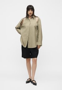 Femme portant une chemise beige surdimensionnée avec des découpes aux épaules, un short noir longueur genoux et des ballerines noires pointues, debout sur fond blanc.