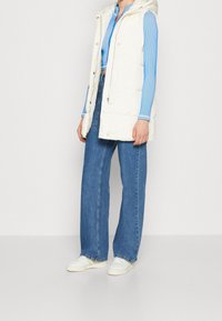 White padded vest with a hood, styled over a blue ribbed long-sleeve top and paired with wide-leg blue jeans and white sneakers.