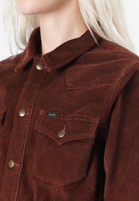 Bruine corduroy shirt met een kraag, voorzien van messing knopen en een borstzak met een blauw label. De textuur is ribbelig en zacht.