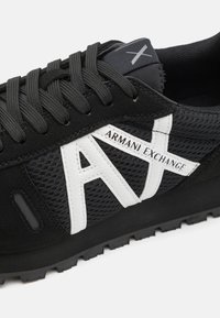 Chaussure de sport noire en tissu mesh, semelle en caoutchouc et logo blanc "Armani Exchange" bien en vue. Design à lacets avec accents et finition texturée.