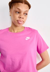 Rožinė medvilninė marškinėliai su balta Nike logo kairėje krūtinės pusėje, apvalia iškirpte ir trumpomis rankovėmis, su lygiu paviršiumi.