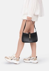Borsa a mano in pelle nera con accessori dorati, caratterizzata da una maniglia superiore e una forma strutturata. Indossata con una camicia bianca e una gonna corta.