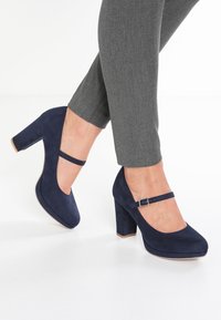 Talons hauts en daim bleu marine avec un bout arrondi et une bride à la cheville. Talon carré épais et texture lisse, associés à un pantalon gris bien ajusté.