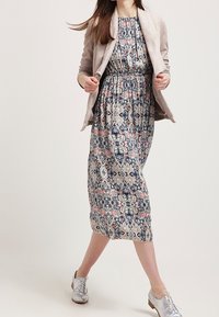 Kvinna i ett mönstrat midiklänning, beige kavaj och glänsande silverfärgade oxfordskor, som går mot en enkel ljus bakgrund.