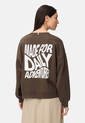 Kobieta z długimi ciemnymi włosami, ubrana w brązową bluzę z białym napisem "MADE FOR DAILY ADVENTURES" na plecach oraz beżową spódnicę.