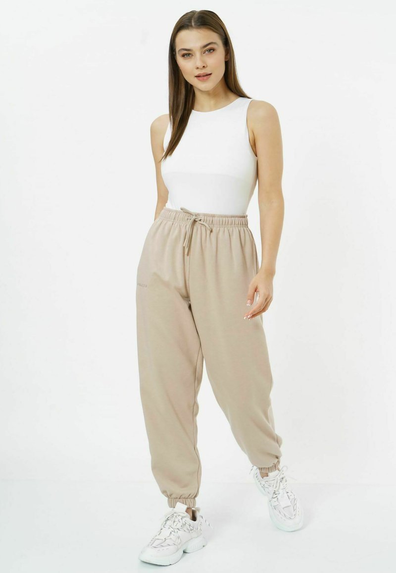 Beige, schmal zulaufende Jogginghosen mit elastischem Bund und Bündchen, kombiniert mit einem figurbetonten weißen Tanktop und weißen Sneakern.