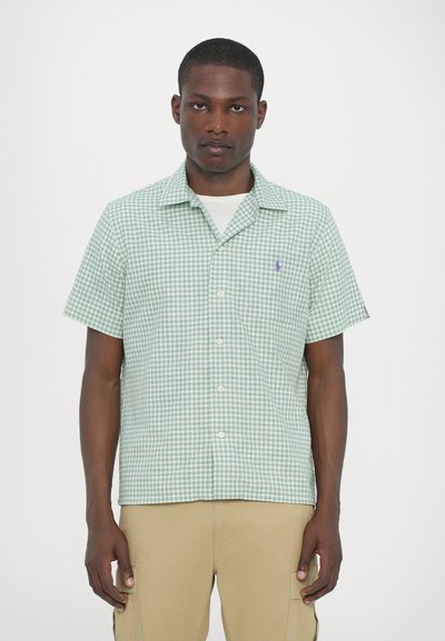 Camisa de cuadros verde y blanca de manga corta, con cuello, de botonadura frontal y un pequeño logo morado en el pecho. Tela ligera.