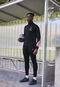Musta urheiluasu, jossa on vaaleanpunaisia yksityiskohtia, yhteensopivat mustat lenkkarit, pitelee mustaa Nike-jalkapalloa metallista taustaa vasten.