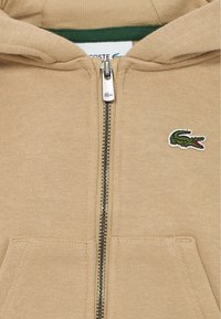 Bežinė zip-up megztinė su žalia vidine pamušalu, kuris turi mažą Lacoste krokodilo logotipą ant krūtinės ir dvi priekinės kišenes.