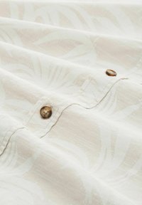 Heller Creme-Stoff mit dezenten Blattmustern, versehen mit braunen Knöpfen entlang der Naht. Weiche Textur mit glatter Oberfläche.